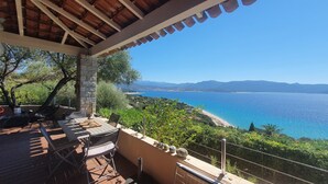 An outdoor patio with stone pillars, a pergola, and a table with chairs. It overlooks a coastal landscape with a clear blue sea and distant mountains.