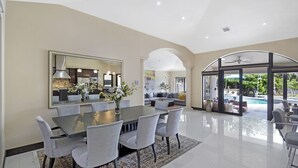 Un comedor amplio con una mesa y sillas grandes, una cocina moderna visible a través de un separador de vidrio, y vista a un área de piscina por un arco.