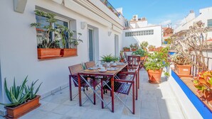 Un pati amb una taula de fusta preparada per dinar, envoltat de plantes en testos i un edifici blanc amb finestres i un balcó.
