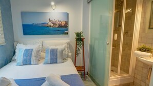 Habitació d'hotel amb llit, dutxa, una planta en test i un quadre d'una ciutat costanera.