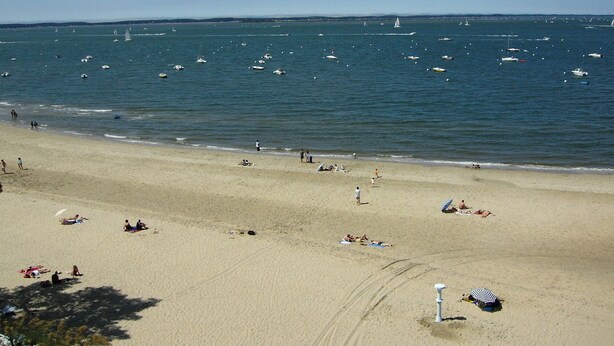 A beach with people sunbathing and swimming, boats on the water, and a clear sky.