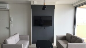 A modern living room with a flat-screen TV mounted on a dark wall, two grey sofas, a wooden coffee table, and a dining table with chairs.