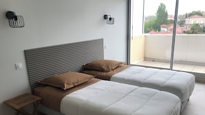A bedroom with two beds, a small wooden table, and a window with a view of buildings.