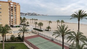 En ejendom ved stranden med tennisbane, palmer og udsigt over havet og fjerne bygninger.