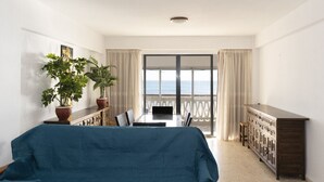 Et værelse med seng, et bord med stole, en potteplante, et træskab og havudsigt fra en balkon.