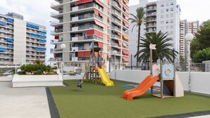 Een speelplaats met glijbanen en klimconstructies, omringd door woongebouwen en palmbomen.