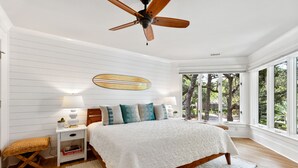 Un dormitorio con cama de madera, paredes blancas, ventilador de techo, ventana grande con vista a árboles y una mesita de noche con lámpara.