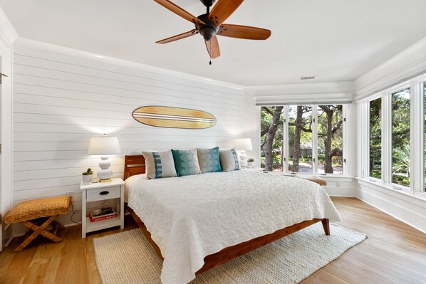 Un dormitorio con cama de madera, paredes blancas, ventilador de techo, ventana grande con vista a árboles y una mesita de noche con lámpara.