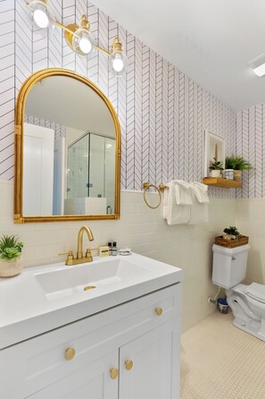 Une salle de bain avec un meuble-lavabo blanc, des accessoires dorés, un grand miroir et un papier peint à motifs.