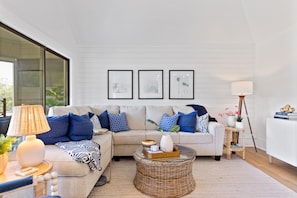 A modern living room with a sectional sofa, a round wicker coffee table, and a side table with a lamp. There are framed pictures on the wall and a potted plant on the floor.