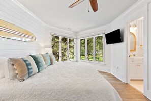 Une chambre spacieuse avec un grand lit, un ventilateur de plafond, un téléviseur fixé au mur et une vue sur l’extérieur grâce à de grandes fenêtres.