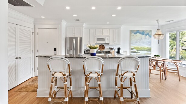 Una cocina moderna con muebles blancos, una isla central con taburetes, electrodomésticos de acero inoxidable y un amplio ventanal con vistas a zonas verdes.