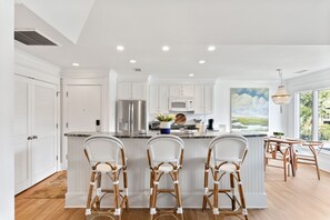 A modern kitchen with white cabinetry, a central island with bar stools, stainless steel appliances, and a large window with a view of greenery.