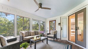 Habitación iluminada por el sol con muebles de mimbre, ventilador de techo y grandes ventanas con vistas a árboles.
