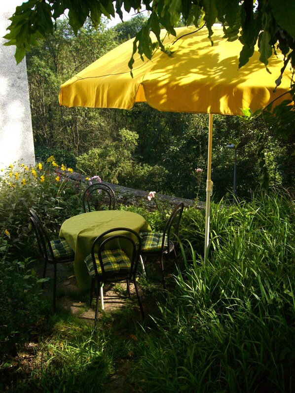 Eine Terrasse im Freien mit Tisch und Stühlen unter einem gelben Sonnenschirm, umgeben von viel Grün.