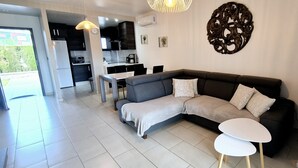 Ein modernes Wohnzimmer mit einem grauen Ecksofa, einem kleinen weißen Tisch und einem Essbereich, im Hintergrund ist eine Küche zu sehen.