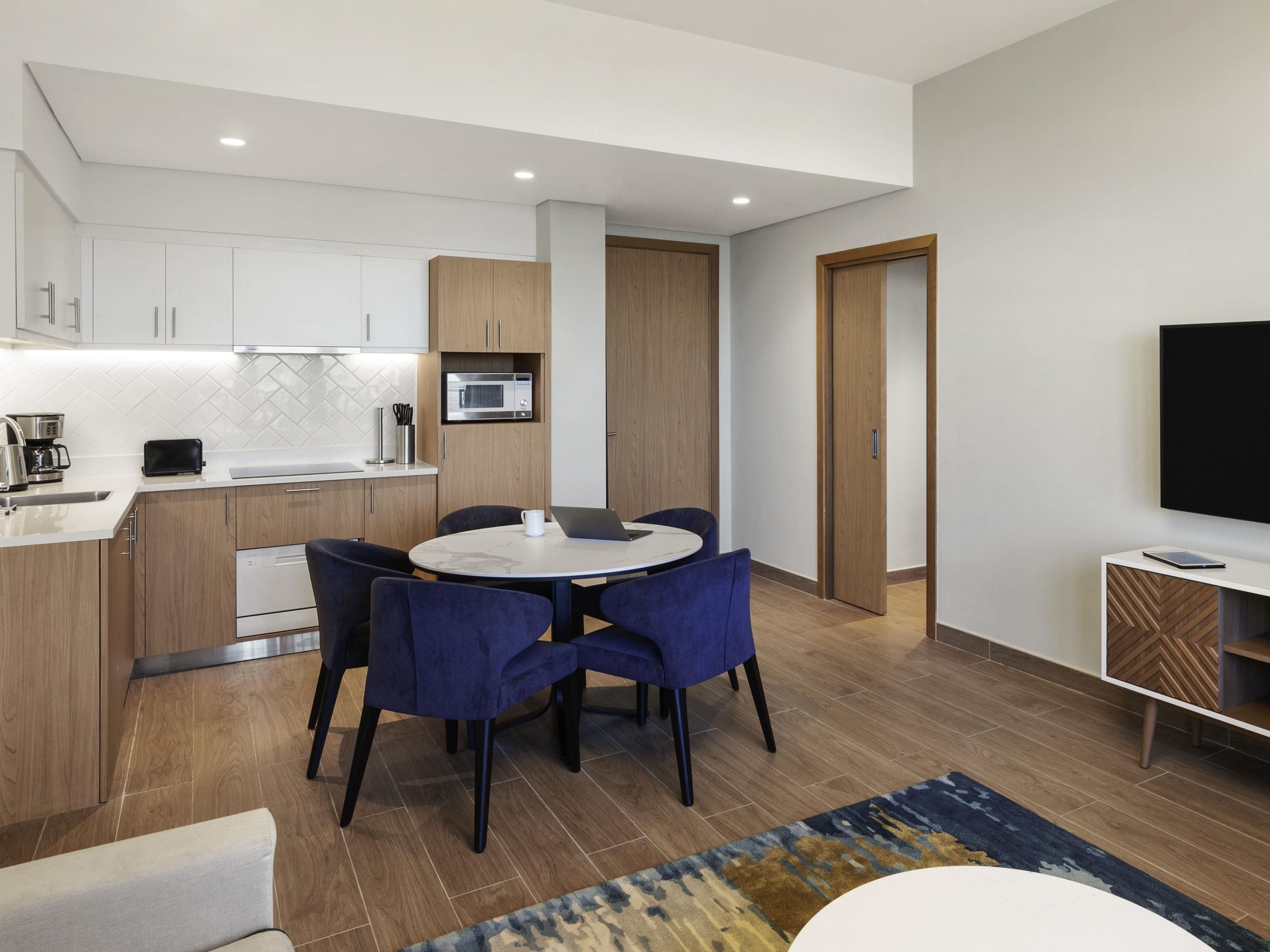 A modern kitchen with white cabinets, a microwave, and a toaster. There is a dining area with a round table and blue chairs.