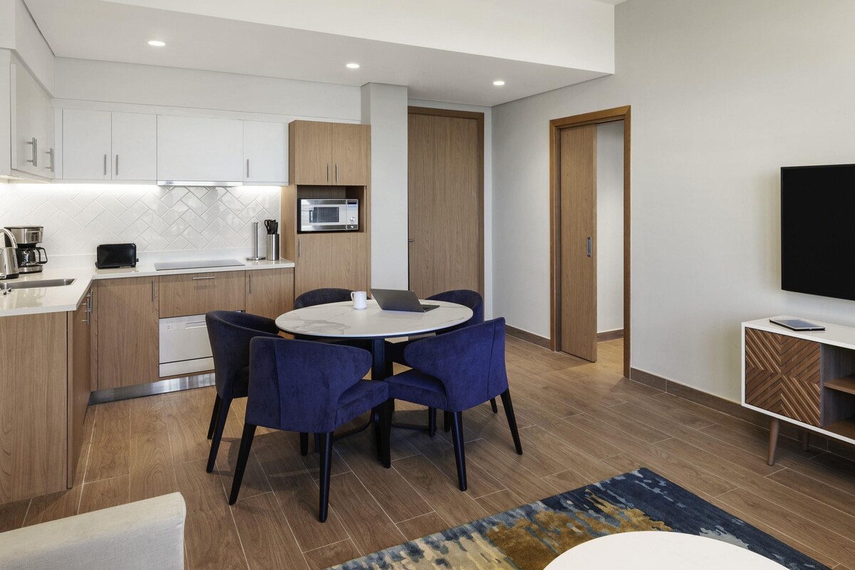 A modern kitchen with white cabinets, a microwave, and a toaster. There is a dining area with a round table and blue chairs.