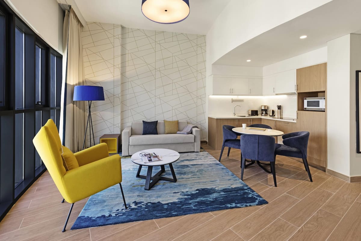 A modern hotel room with a sofa, yellow armchair, dining table, and a kitchen area.