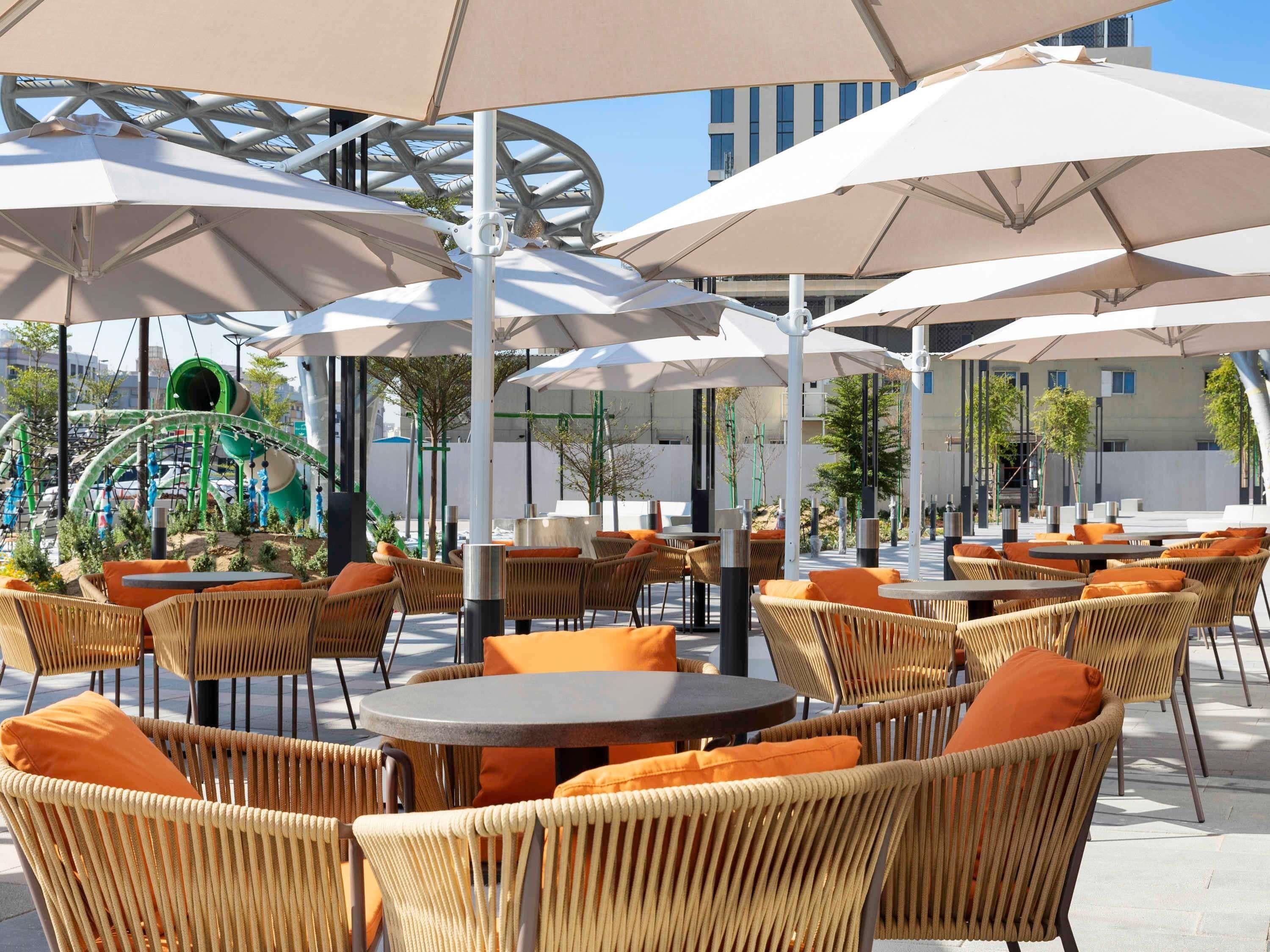 An outdoor seating area with wicker chairs and round tables, shaded by large umbrellas.