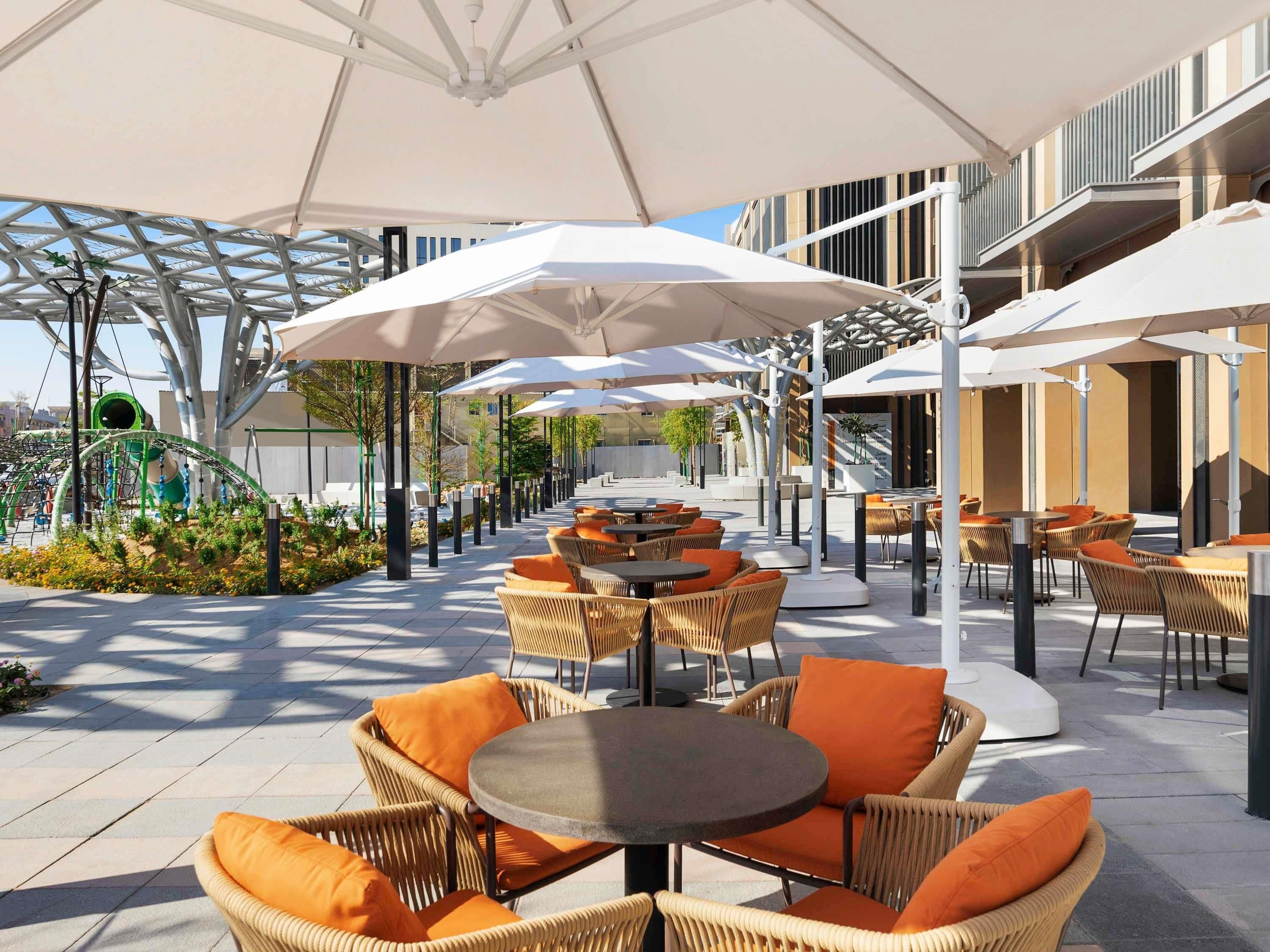 Un espace extérieur aménagé avec des chaises et des tables en osier, protégé par de grands parasols, et un bâtiment moderne en arrière-plan.