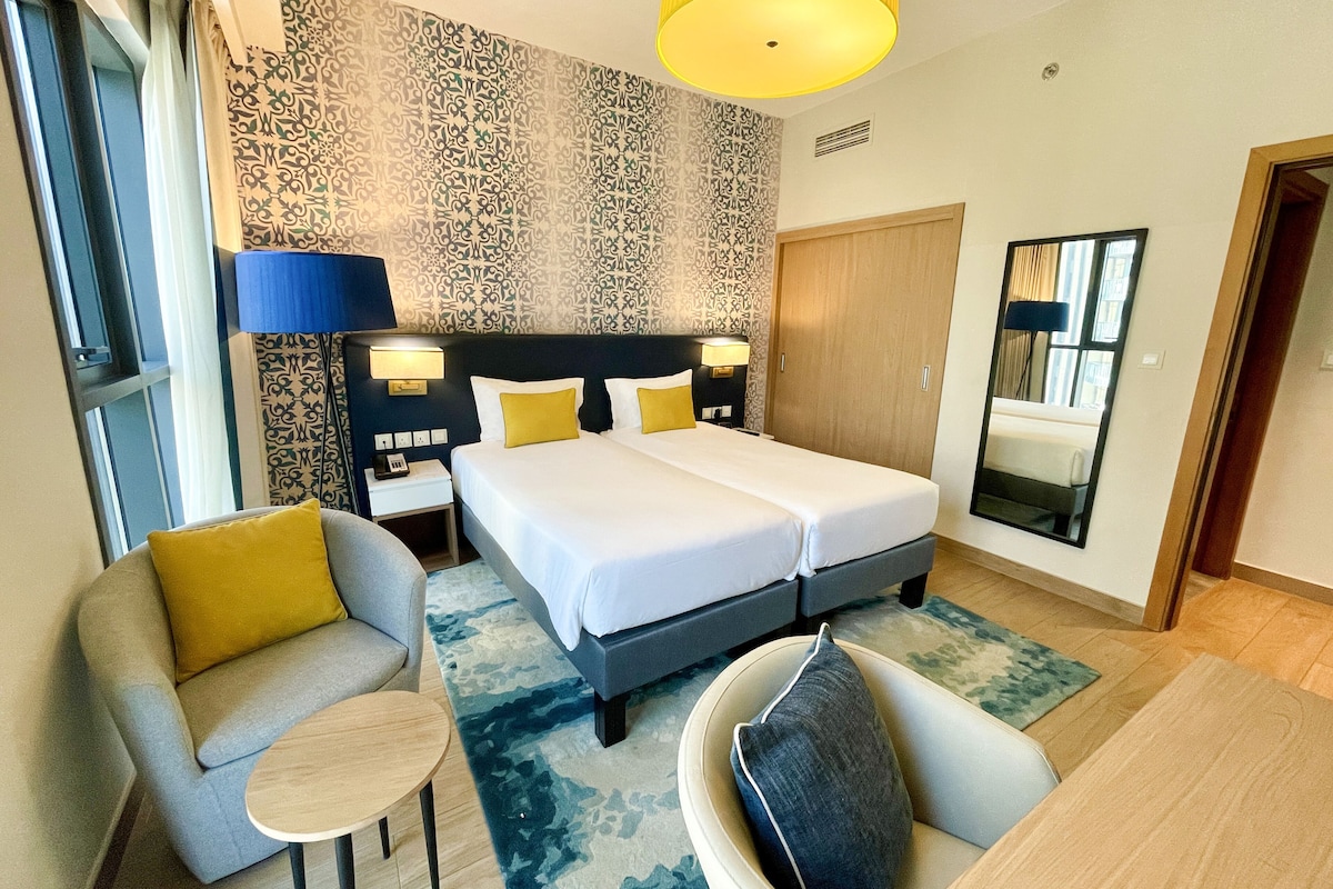 Habitación de hotel con cama doble, dos mesitas de noche con lámparas, un sofá, una mesa redonda pequeña y un espejo.