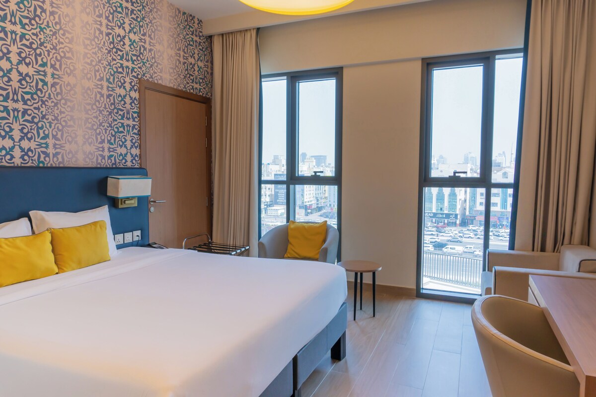 Habitación de hotel con una cama grande, un escritorio, una silla y vistas de la ciudad a través de amplias ventanas.