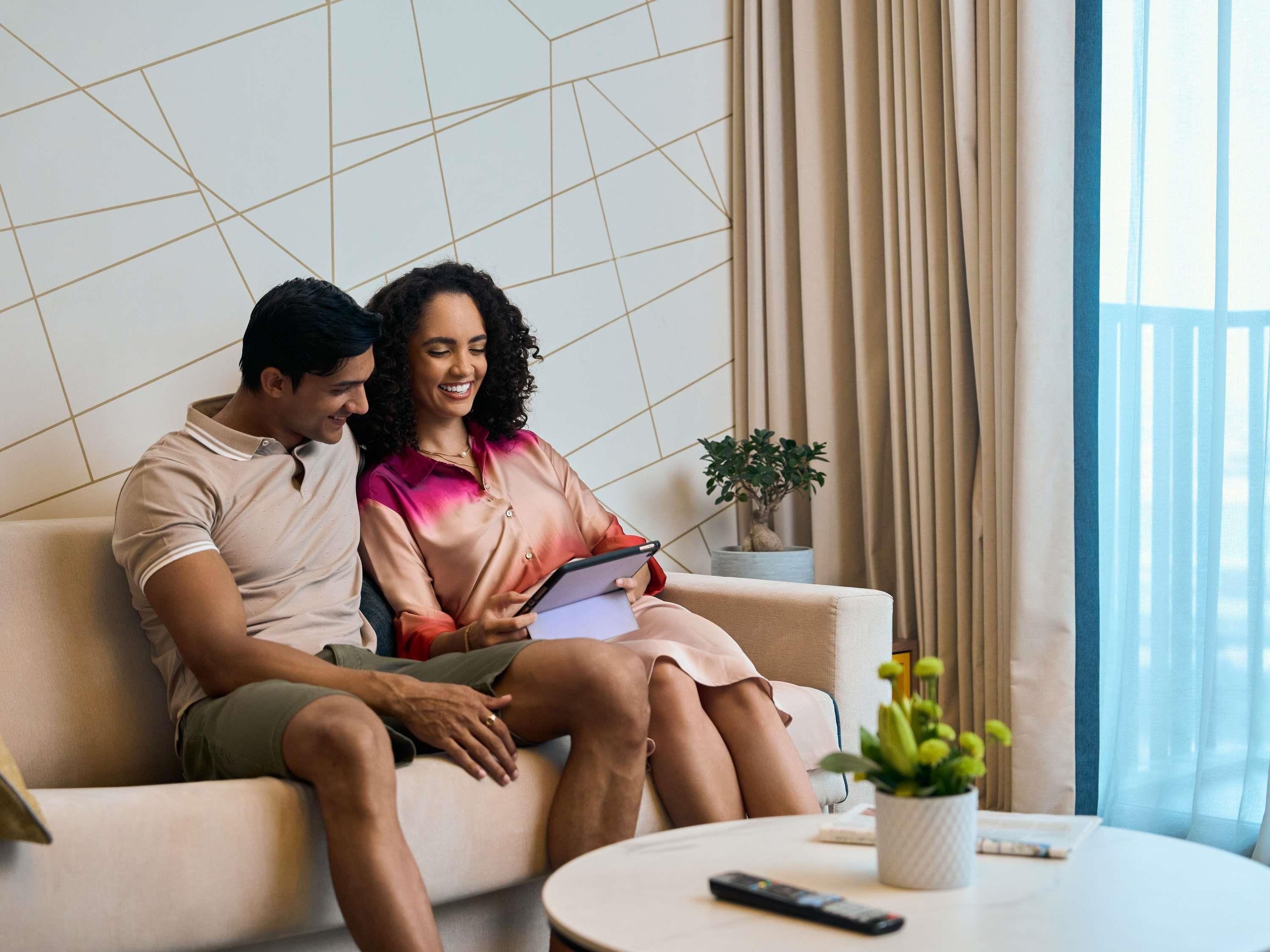 Un couple est assis sur un canapé dans un salon moderne, avec un mur décoré de motifs géométriques, une plante en pot et une télécommande posée sur une table d’appoint.