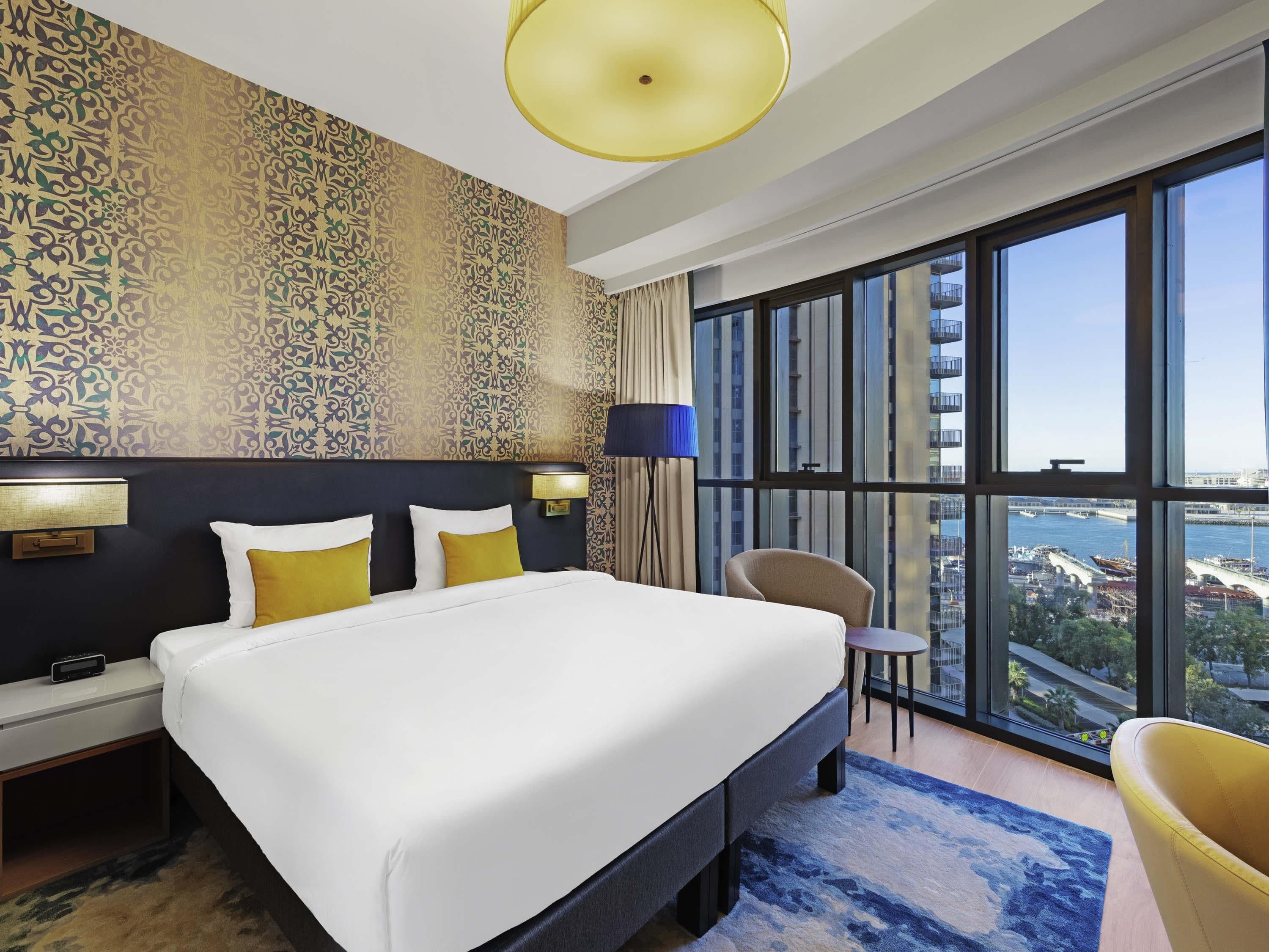 A hotel room with a large bed, a desk, a chair, a large window with a city view, and patterned wallpaper.