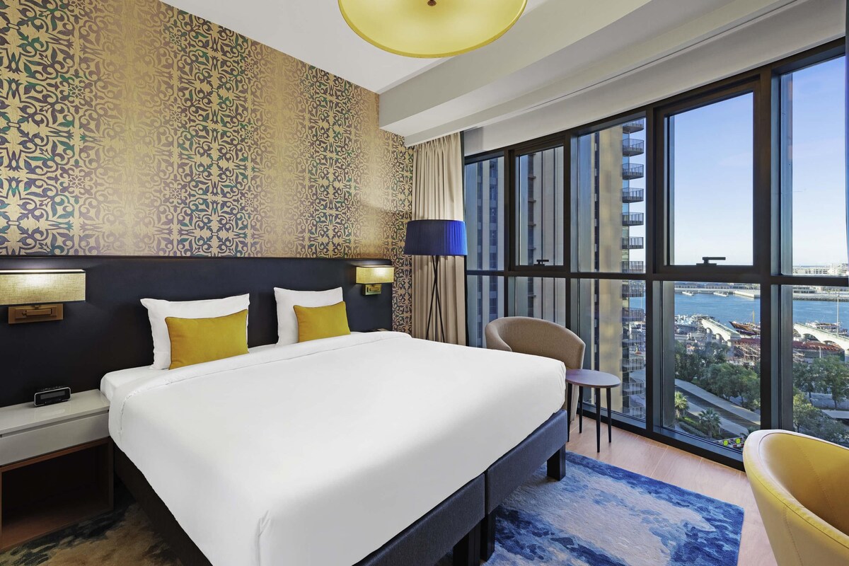 A hotel room with a large bed, a desk, a chair, a large window with a city view, and patterned wallpaper.
