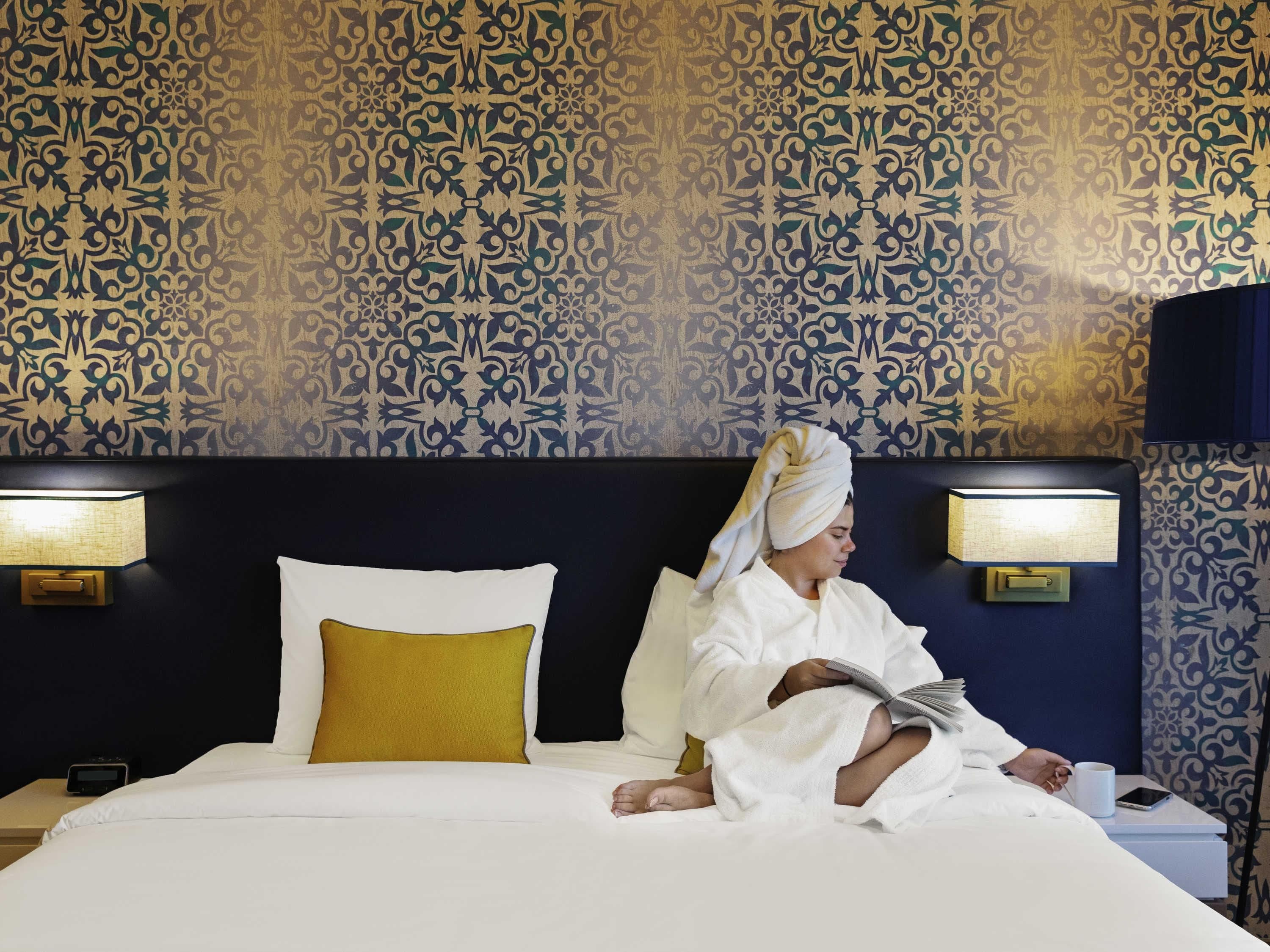 A hotel room with a patterned wallpaper, a bed with white and yellow pillows, and bedside lamps.