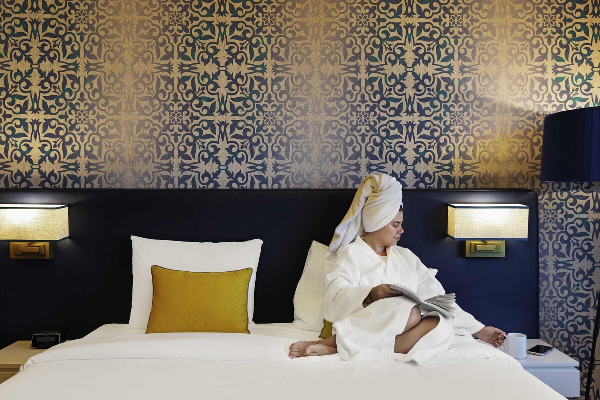 A hotel room with a patterned wallpaper, a bed with white and yellow pillows, and bedside lamps.