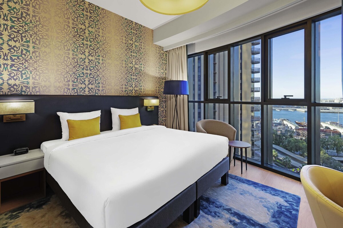 A hotel room with a large bed, a desk, a chair, a large window with a city view, and patterned wallpaper.