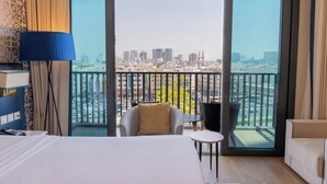 Habitación de hotel con una cama grande, una silla, una mesita y vista a un paisaje urbano desde un balcón.