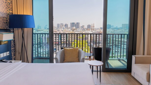 Habitación de hotel con una cama grande, una silla, una mesita y vista a un paisaje urbano desde un balcón.