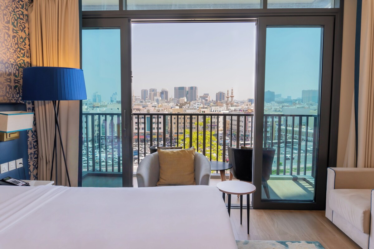 Habitación de hotel con una cama grande, una silla, una mesita y vista a un paisaje urbano desde un balcón.