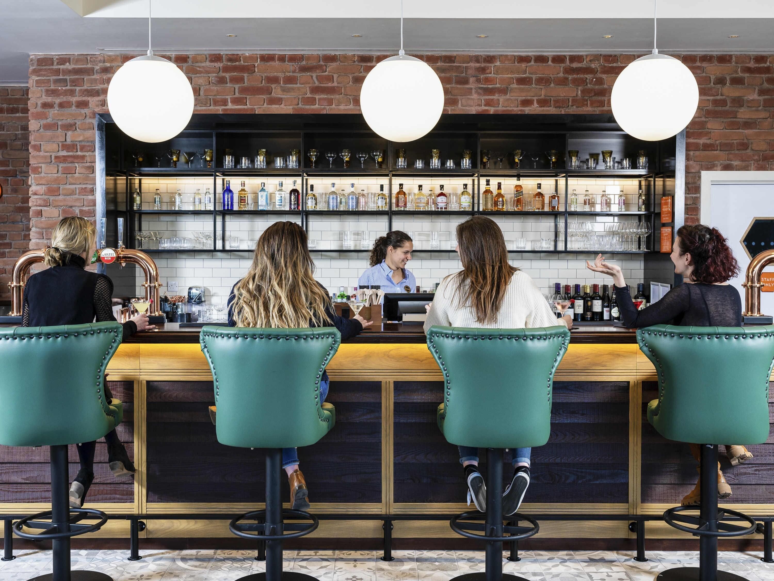A bar area with a bartender, four bar stools, and a brick wall with shelves of bottles.
