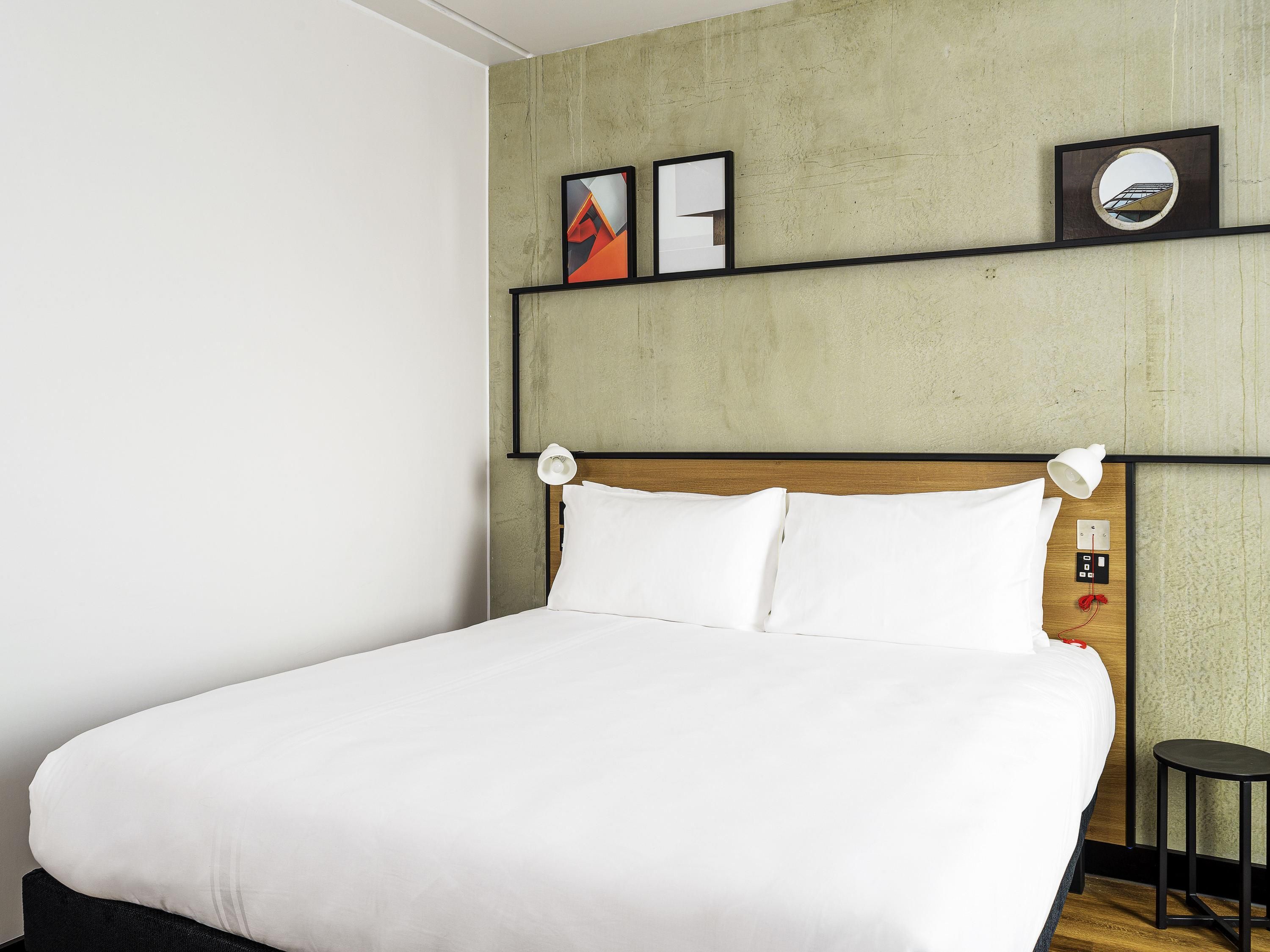 Una cama bien hecha con sábanas blancas en una habitación con pared de hormigón y cabecero de madera.