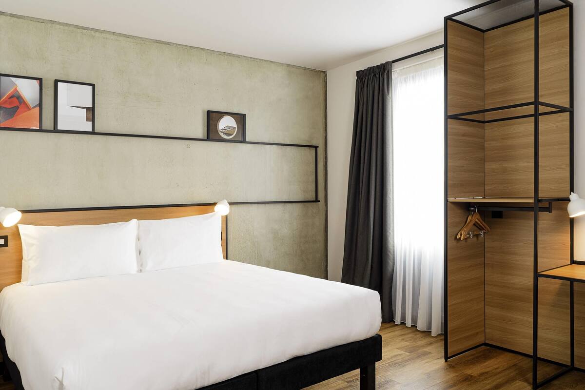 A neatly arranged hotel room with a large bed, a wooden wardrobe, and a wall-mounted shelf with pictures.