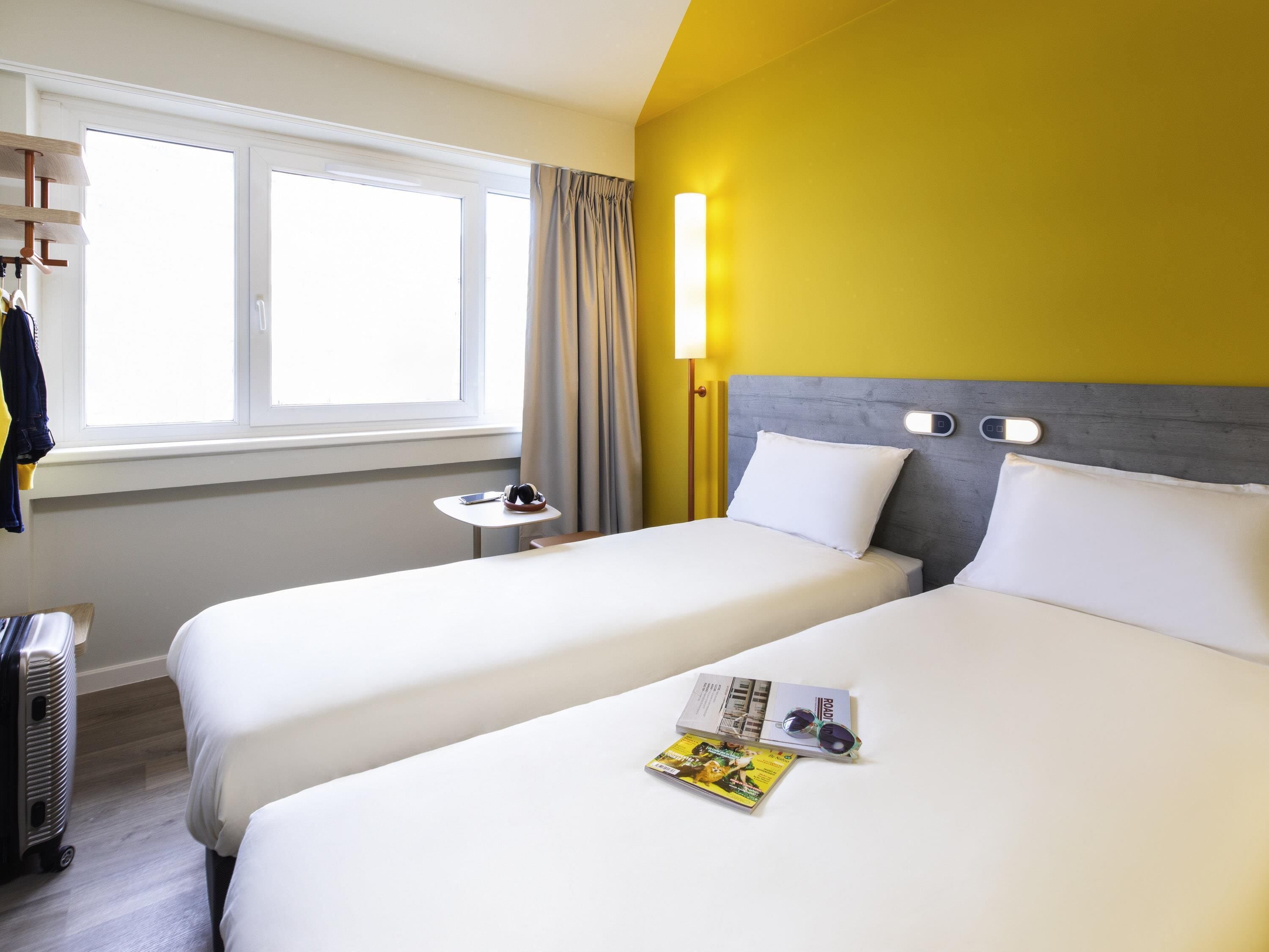 Een hotelkamer met twee bedden, een raam met gordijnen, een nachtkastje met een lamp en een koffer.