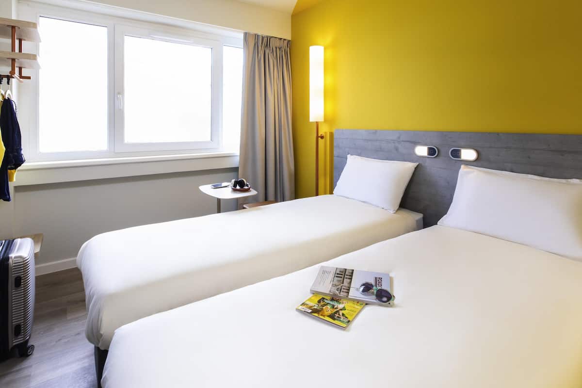 Een hotelkamer met twee bedden, een raam met gordijnen, een nachtkastje met een lamp en een koffer.