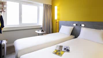 A hotel room with two beds, a window with curtains, a bedside table with a lamp, and a suitcase.