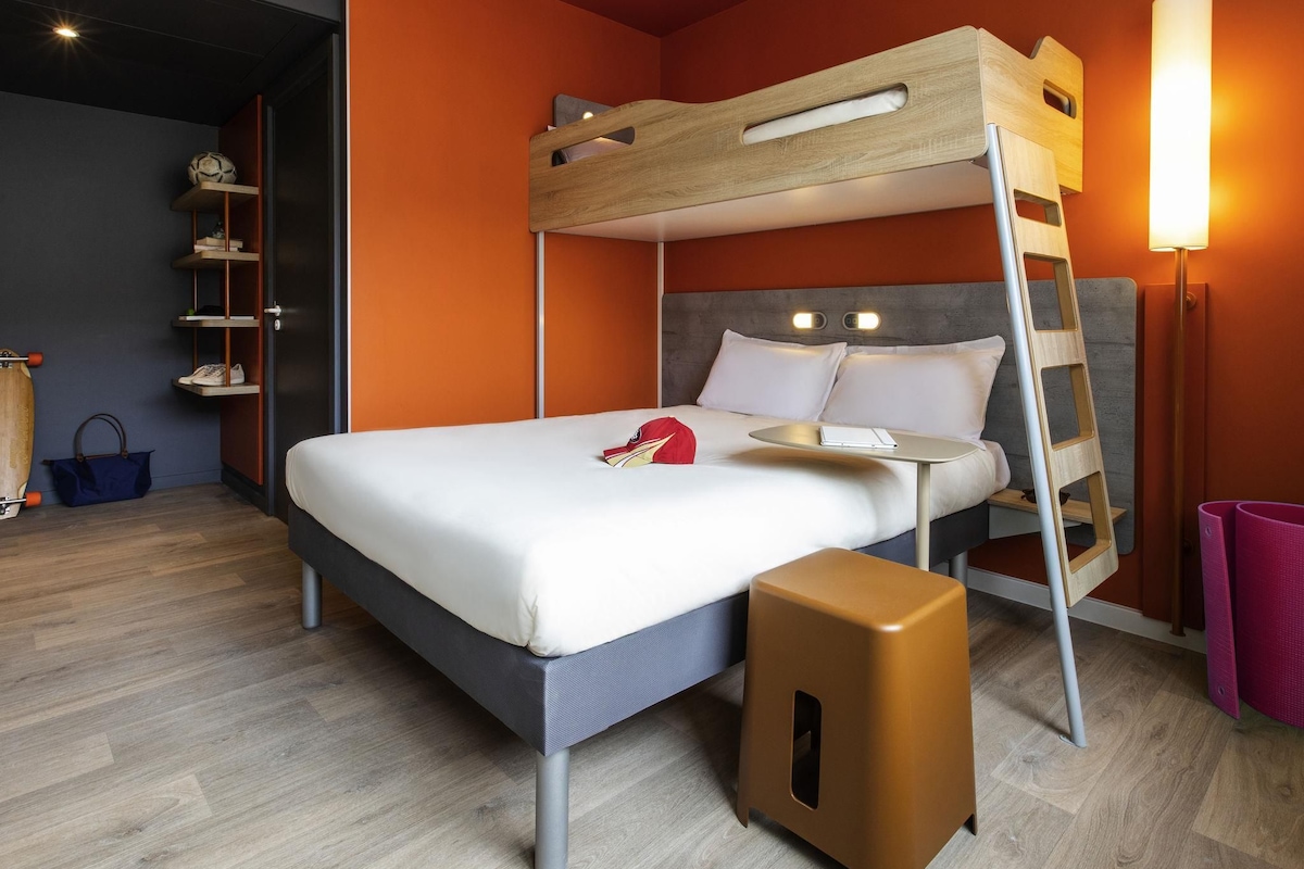 Un lit superposé avec un bureau et une chaise, un petit tabouret et une lampe fixée au mur.