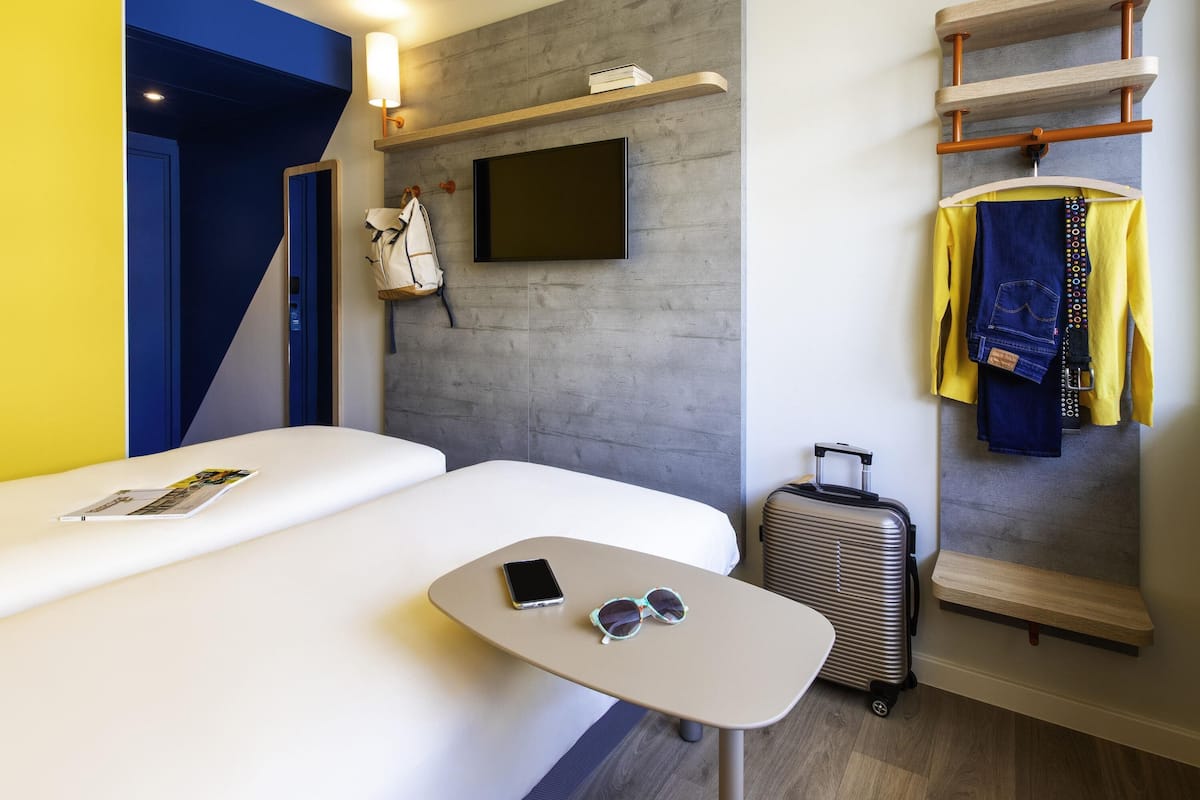 A hotel room with a bed, a small table with a phone and sunglasses, a suitcase, a coat rack with clothes, and a mounted TV.