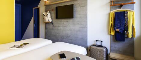 Et hotelværelse med en seng, et lille bord med telefon og solbriller, en kuffert, et knagered med tøj og et ophængt tv.