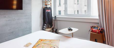 A hotel room with a bed, a small table, a guitar, a helmet, and a board game.