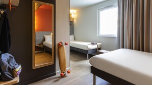 Une chambre d’hôtel avec un lit, un canapé, une petite table, un miroir et un skateboard appuyé contre le mur.