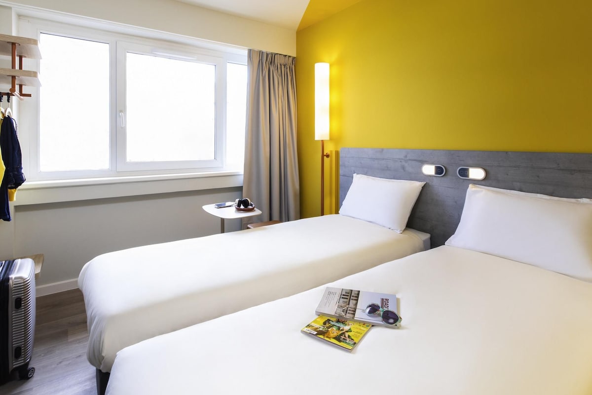 Une chambre d’hôtel avec deux lits, une fenêtre avec des rideaux, une table de chevet avec une lampe et un magazine sur le lit.