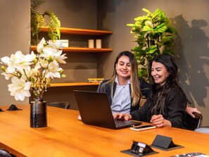 Two individuals at a wooden table with a laptop, a vase of white flowers, and a plant in the background. - Novotel Criciuma (Criciuma)
