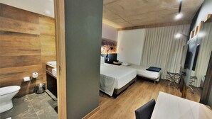 A modern hotel room with a wooden floor, a bed with a white bedspread, a small bedside table with a lamp, a TV mounted on the wall, and a window with curtains. - Novotel Criciuma (Criciuma)